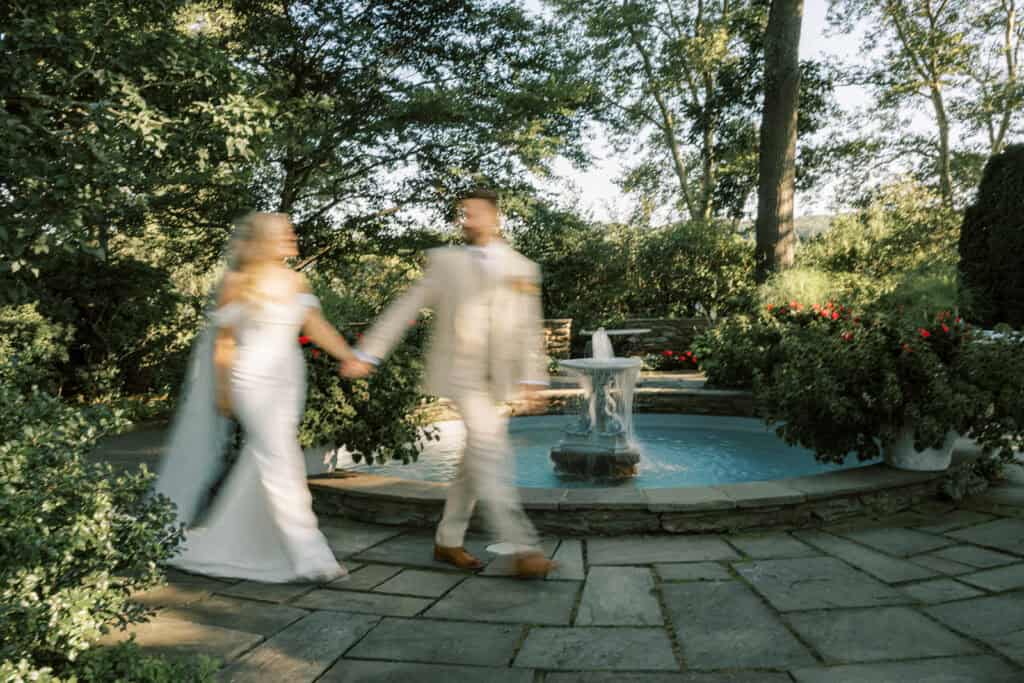 Motion blur photo of bride and groom walking hand in hand past stone fountain in Drumore Estate garden at Lancaster PA summer wedding