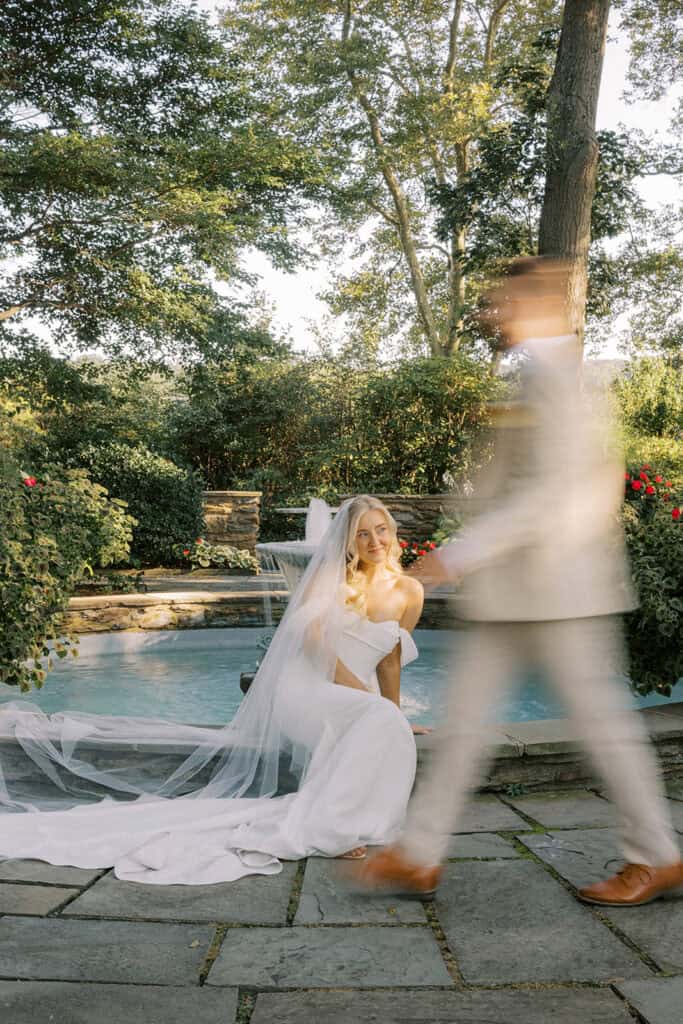 Artistic motion blur of groom walking toward bride seated poolside in Madi Lane gown and cathedral veil at Drumore Estate summer wedding
