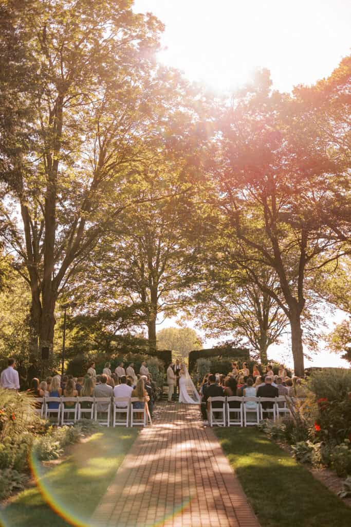 Wide shot of outdoor wedding ceremony at Drumore Estate Lancaster PA at golden hour with guests seated along brick aisle under tall trees