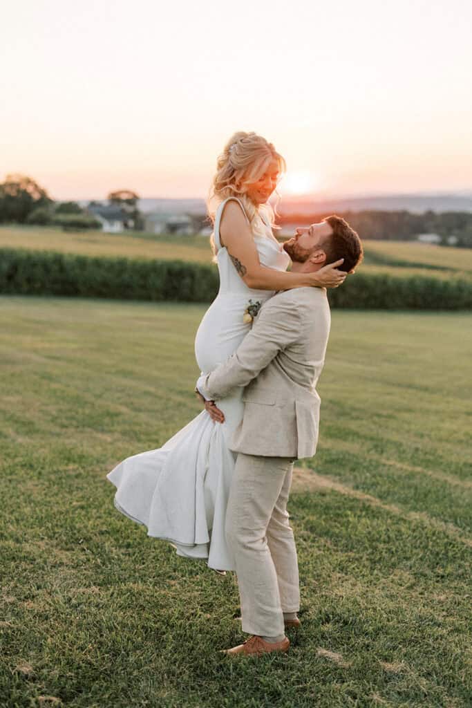 Groom lifting bride in Madi Lane gown at golden hour during summer wedding sunset portraits at Drumore Estate Lancaster PA