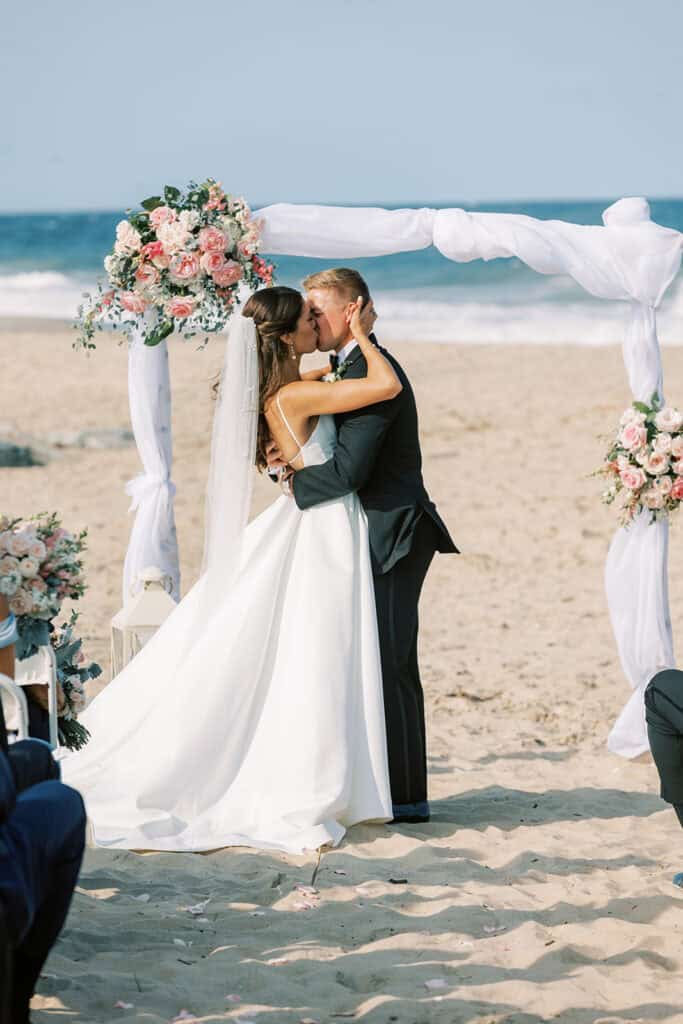Bride and groom's first kiss under blush floral wedding arch on Rehoboth Beach at Henlopen Acres Beach Club, Delaware wedding photographer