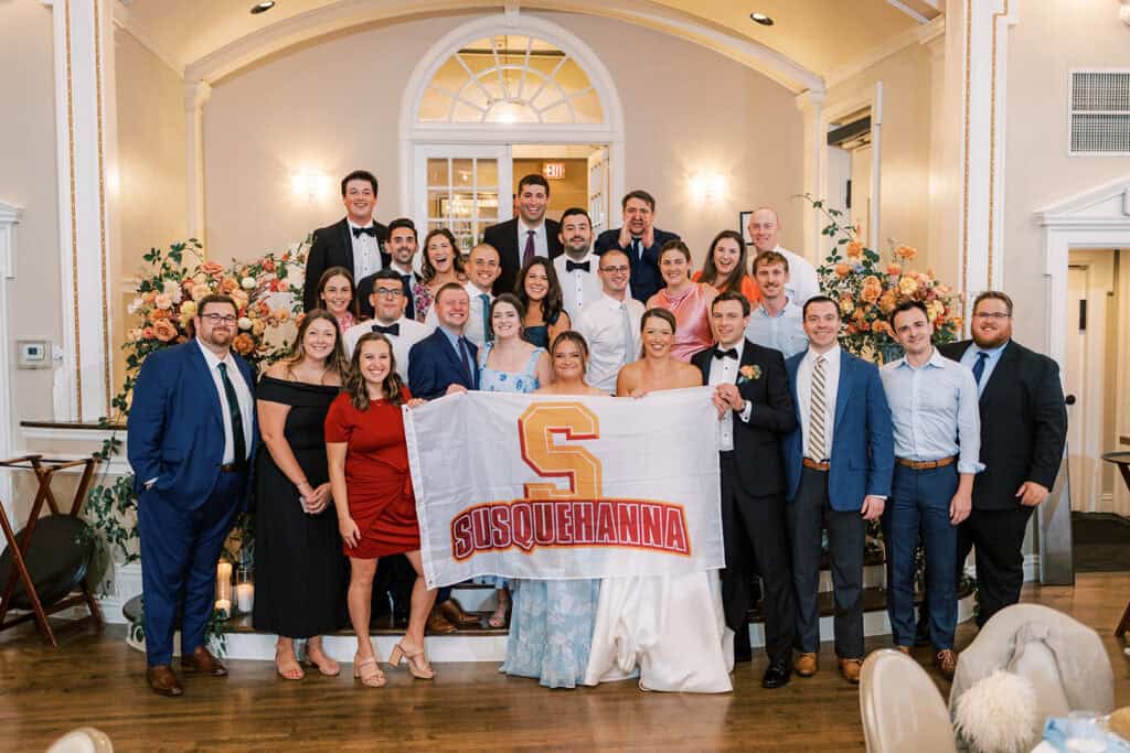 Large group of wedding guests stand with the bride and groom holding a Susquehanna University flag