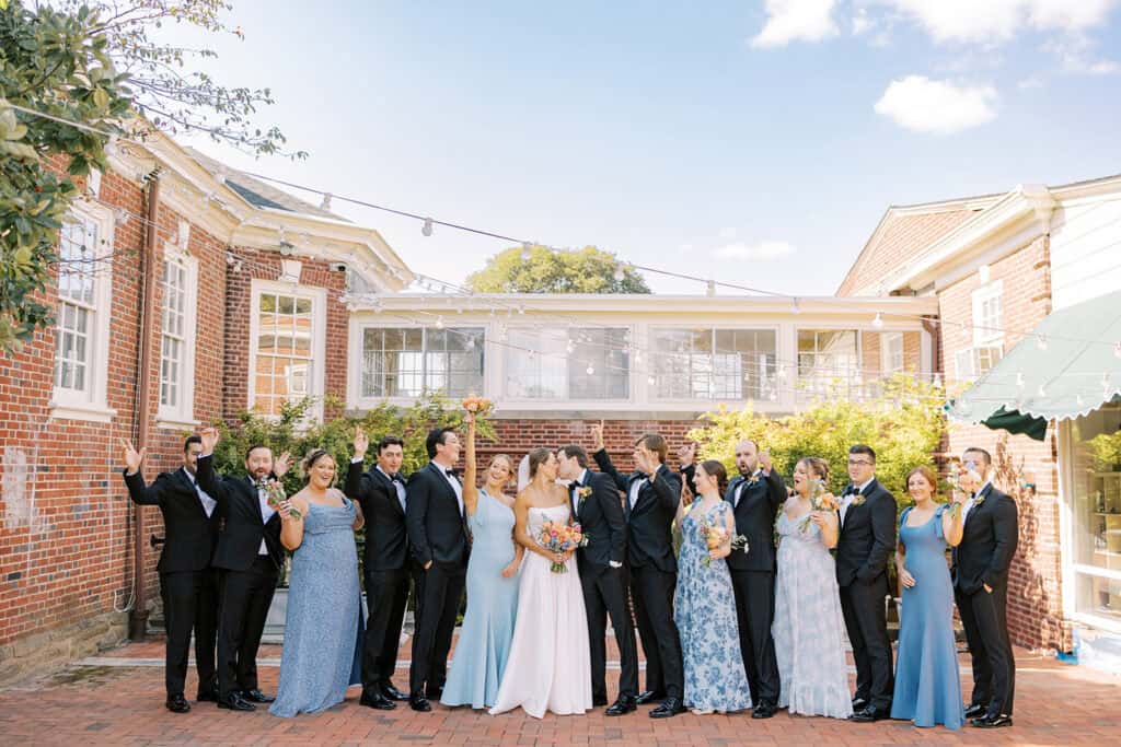 Jena and James kissing at the center of their cheering wedding party in blue dresses and black tuxedos, the brick clubhouse and string lights behind them, at the Philadelphia Cricket Club
