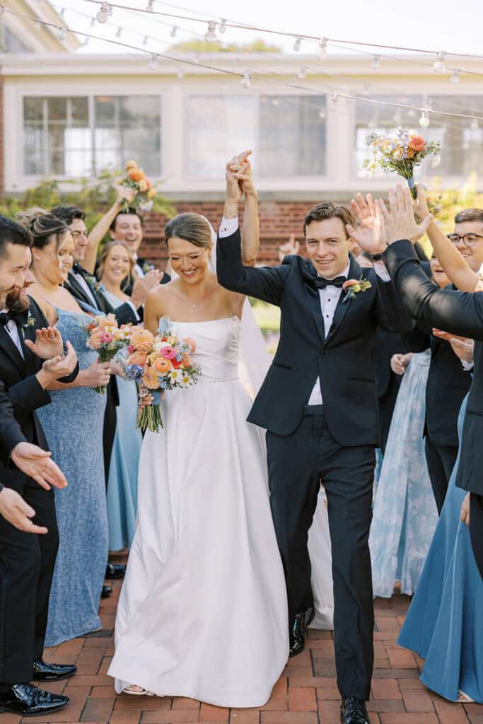 The newlyweds walking hand in hand through their cheering wedding party with arms raised, brick courtyard and string lights overhead, a joyful September Philadelphia Cricket Club wedding moment