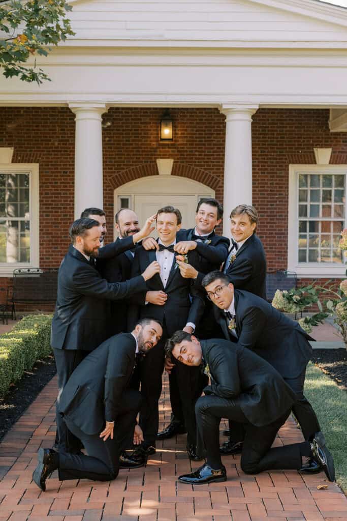 James and his groomsmen in black tuxedos clowning around him in front of the brick Philadelphia Cricket Club clubhouse, an early fall wedding by Samantha Jay Photo