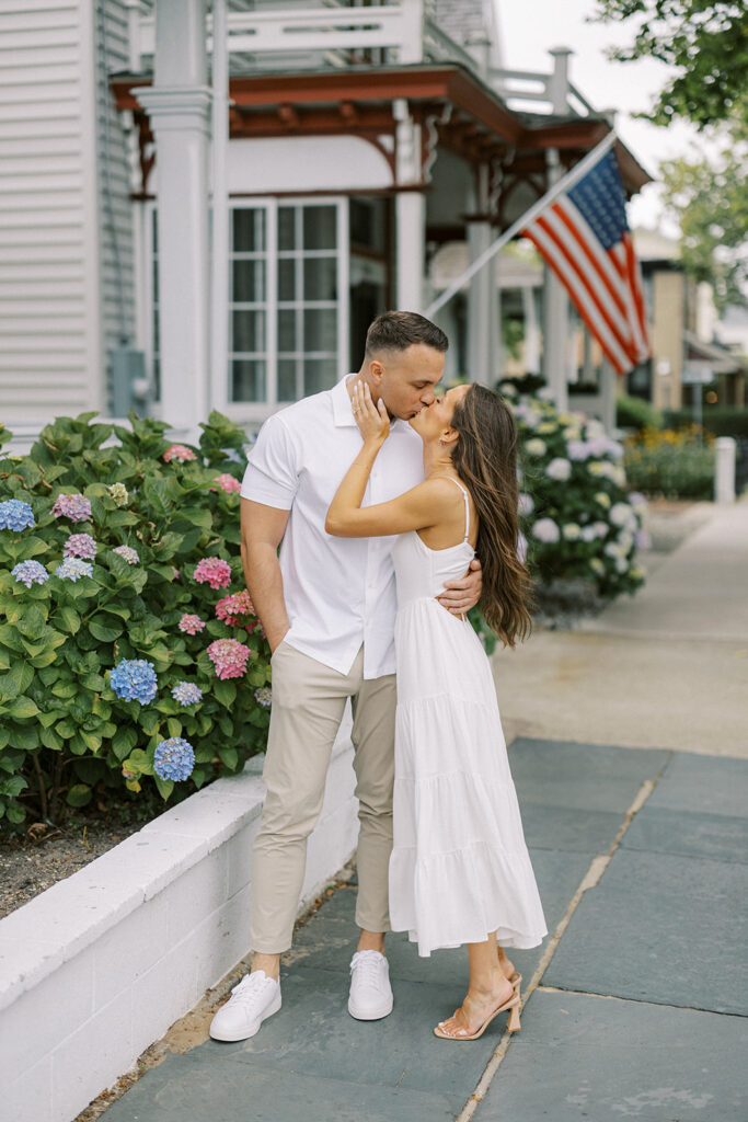Carolyn and Zach kissing on a Cape May sidewalk beside colorful hydrangeas and an American flag on a Victorian porch during their engagement session