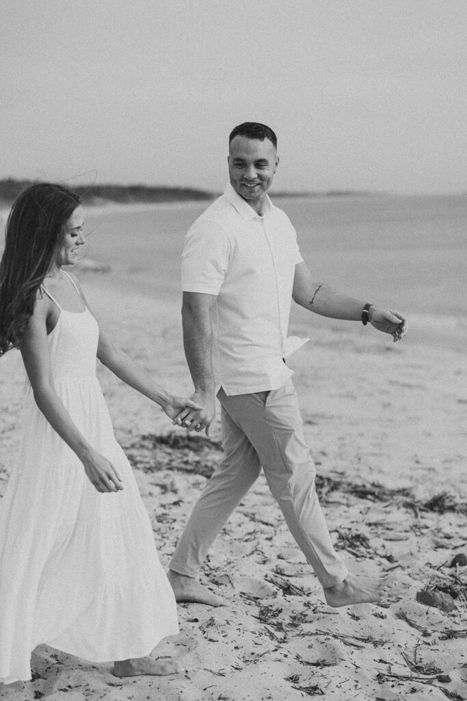 Black-and-white photo of Carolyn and Zach walking hand in hand barefoot along the beach at their Cape May engagement session