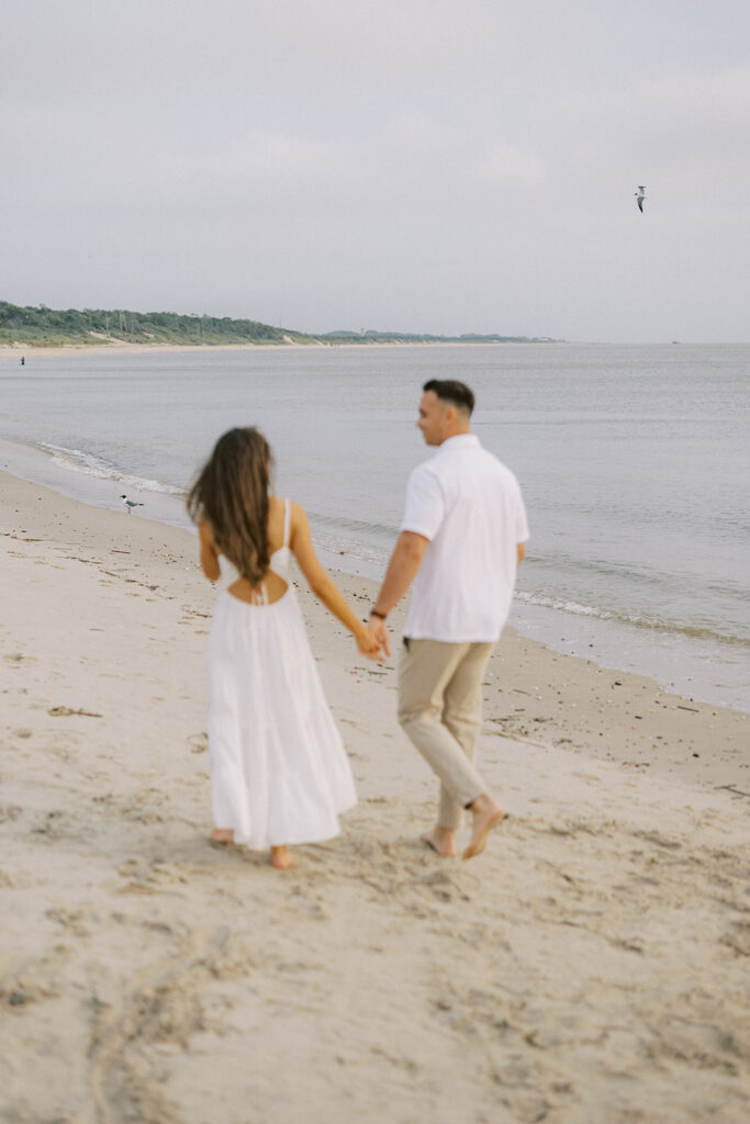 Engaged couple holding hands as they walk toward the bay with a seagull overhead during their Cape May beach engagement session