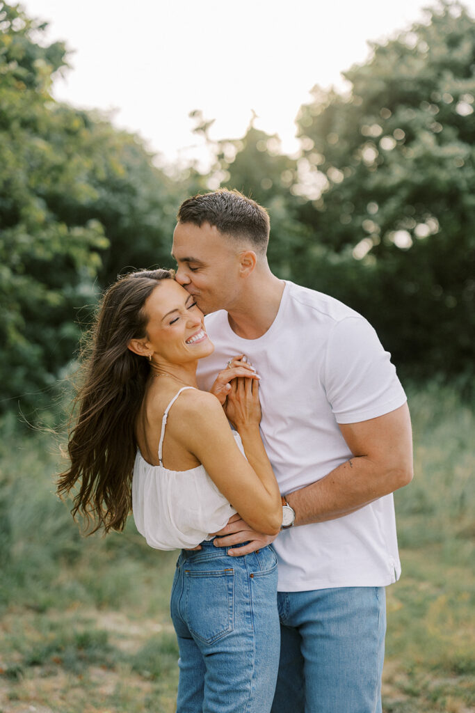 Zach kissing Carolyn's forehead as she laughs in a sunlit field during their golden-hour Cape May engagement session