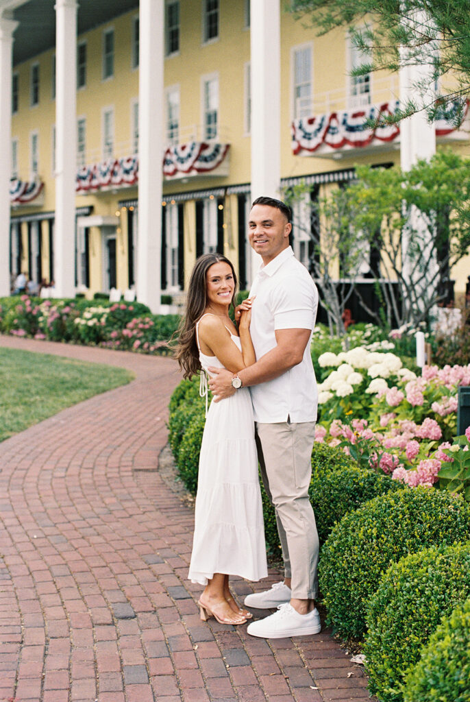 Carolyn and Zach embracing on the brick garden path outside Congress Hall during their Cape May engagement session, surrounded by pink and white hydrangeas