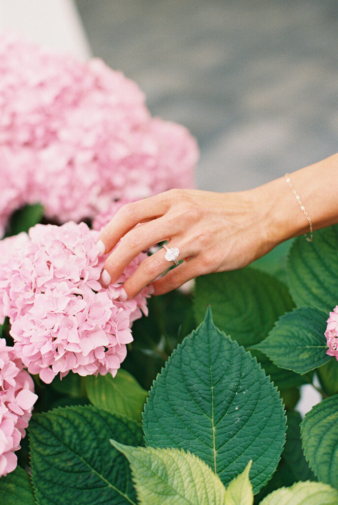 Oval diamond engagement ring detail as the bride-to-be touches a pink hydrangea bloom during her Cape May engagement session