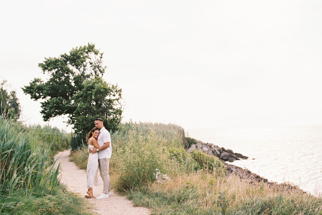Engaged couple embracing on a grassy coastal path overlooking Delaware Bay at their Cape May engagement session