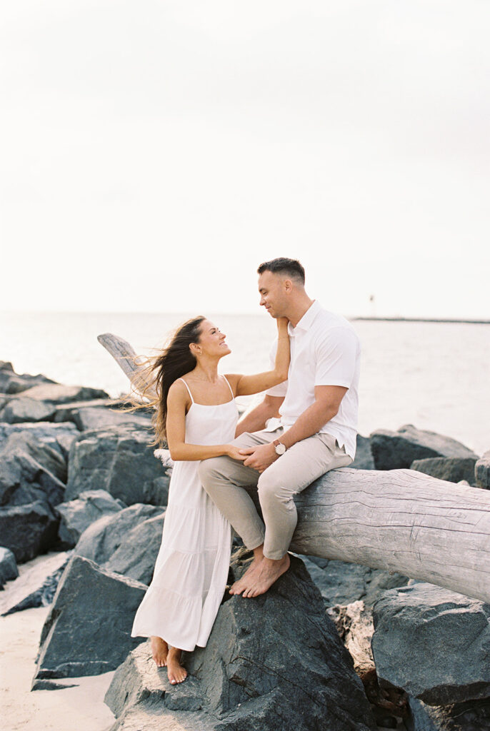 Couple sitting on driftwood among the jetty rocks by the water at their Cape May beach engagement session