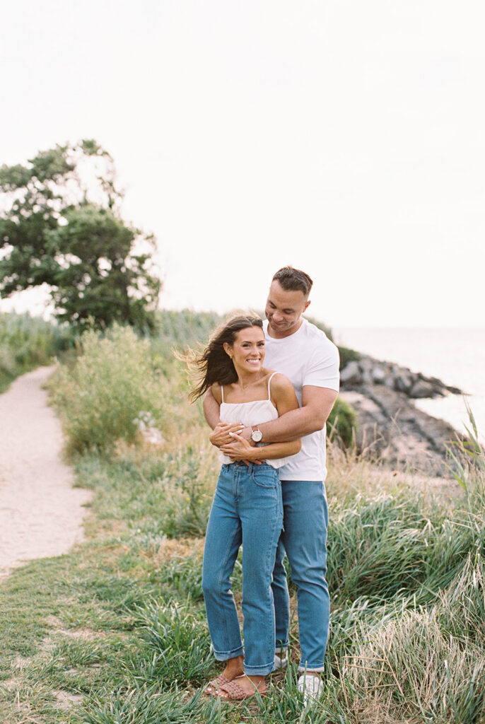 Carolyn and Zach laughing in denim and white tops on a windswept coastal path during their Cape May engagement session