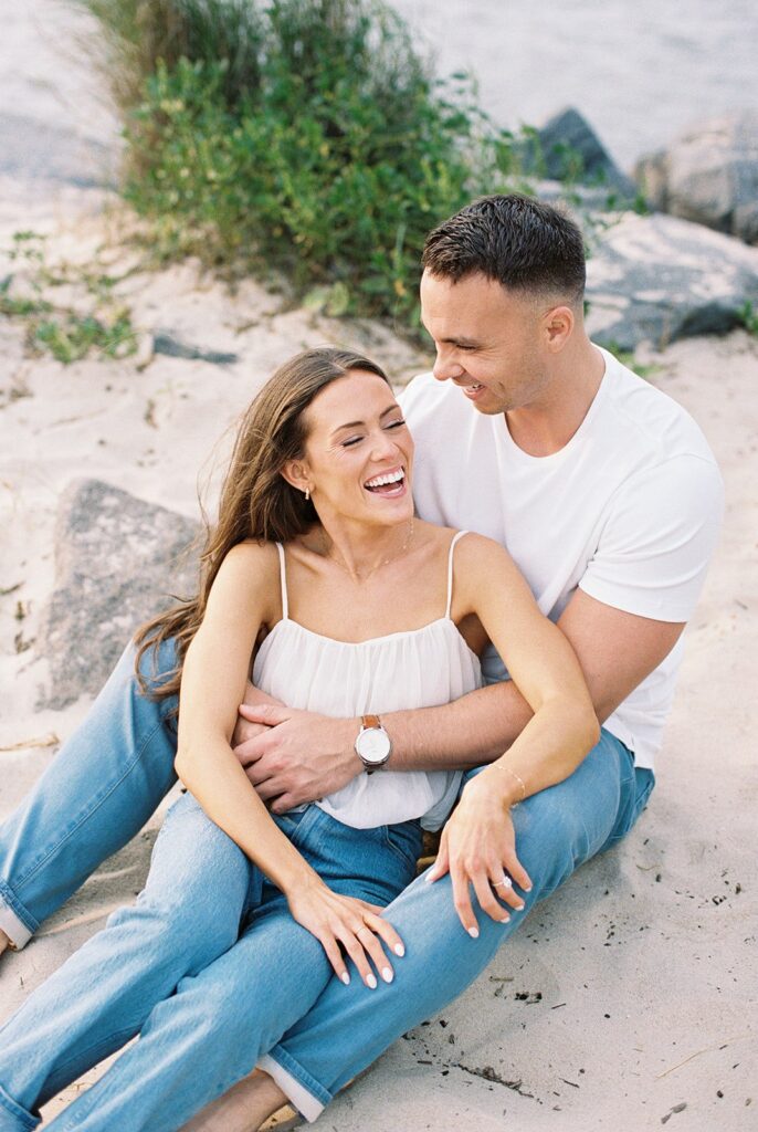 Carolyn and Zach laughing together seated on the beach at golden hour during their Cape May engagement session