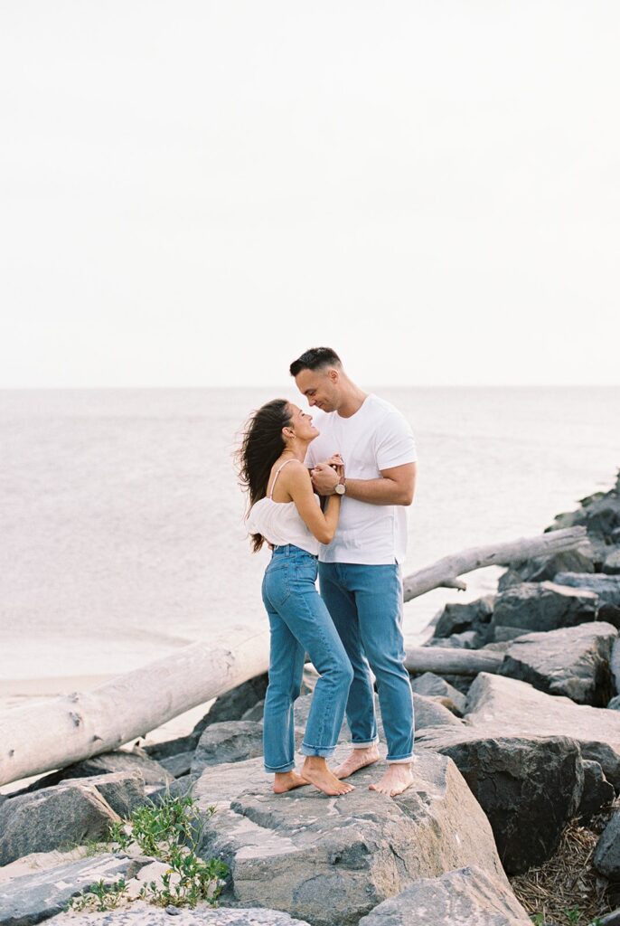 Carolyn and Zach standing barefoot on the jetty rocks, foreheads touching, with the bay behind them at their Cape May engagement session