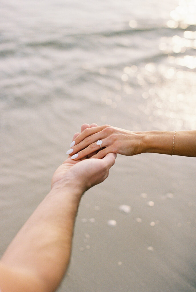 Oval diamond engagement ring detail as the couple's hands meet over the sunlit water at their Cape May beach engagement session