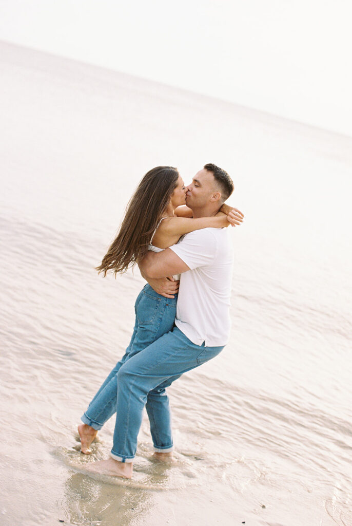 Zach lifting Carolyn for a kiss in the shallow water during their playful Cape May beach engagement session