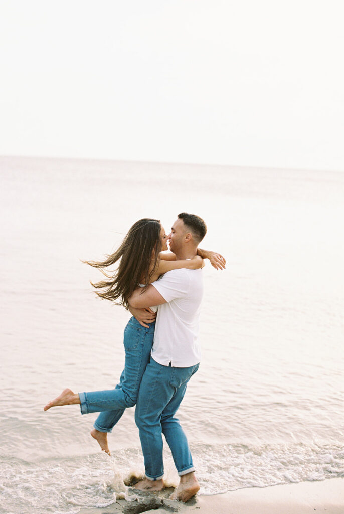 Engaged couple embracing and laughing at the water's edge as Carolyn kicks up her foot at their Cape May engagement session