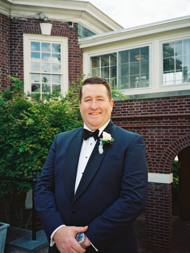 Groom in a navy tuxedo with a white rose boutonniere outside the brick clubhouse, shot on 35mm film