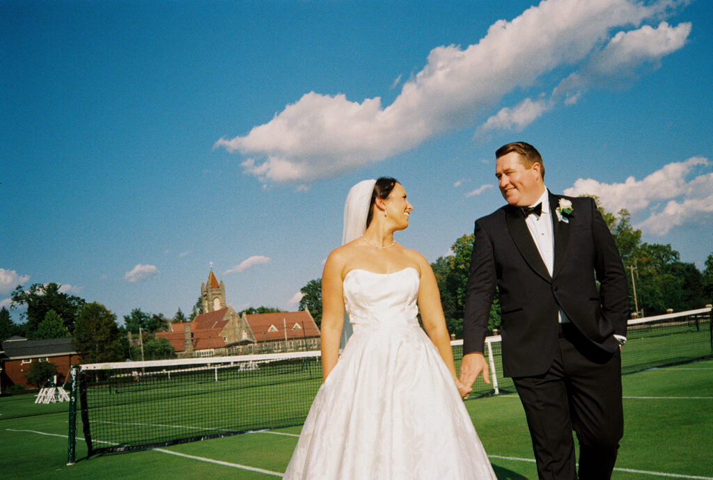 Bride and groom walking hand in hand across the grass tennis courts at the Philadelphia Cricket Club under a blue sky, on 35mm film