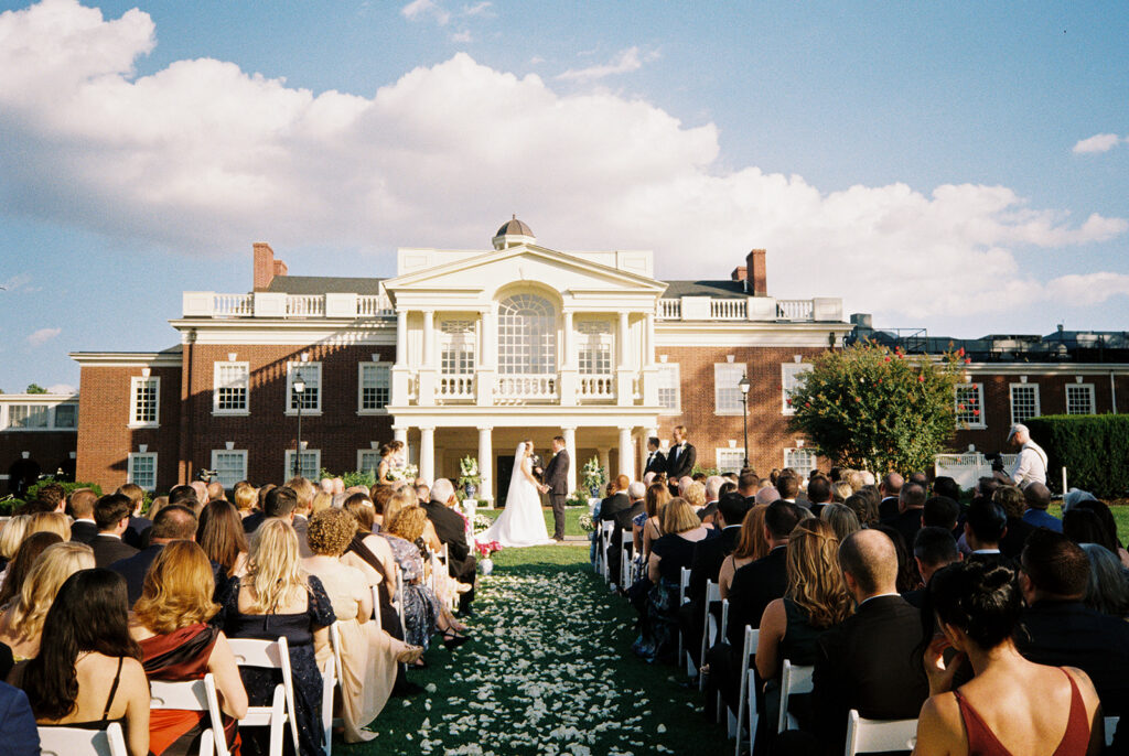 Wide film photo of a blue and white Philadelphia Cricket Club wedding ceremony, guests seated along the petal-lined aisle before the brick clubhouse
