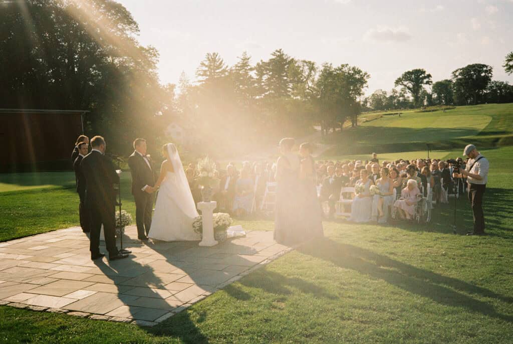 Golden-hour lawn ceremony on film, the couple holding hands as sun flares over guests and the golf course at the Philadelphia Cricket Club