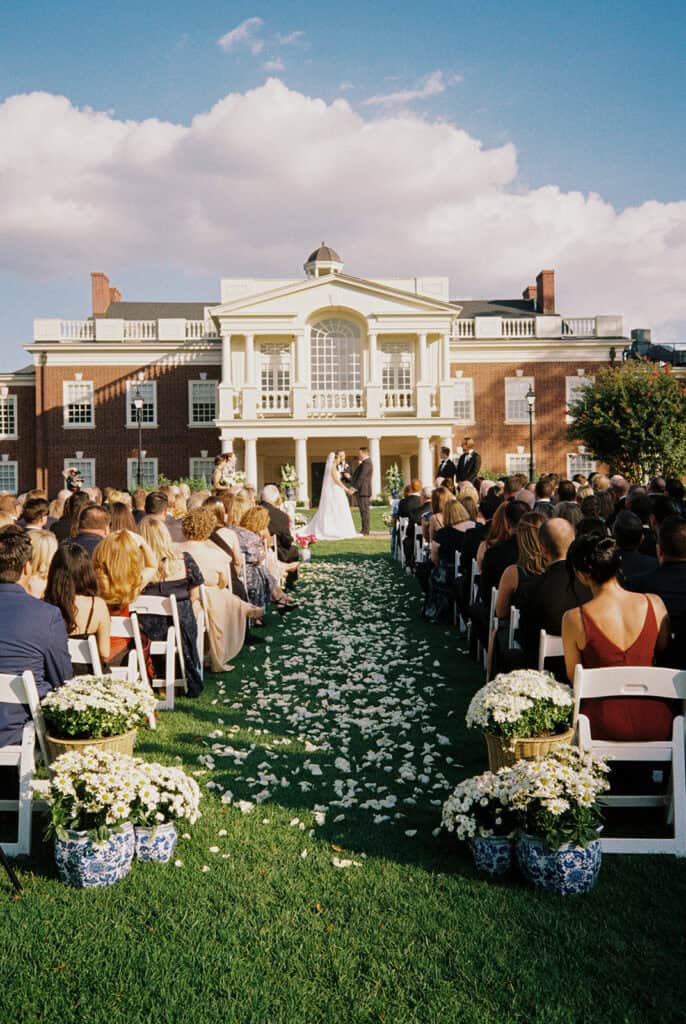 Wide film photo of a blue and white Philadelphia Cricket Club wedding ceremony, blue and white china vases lining the petal-strewn aisle before the brick clubhouse