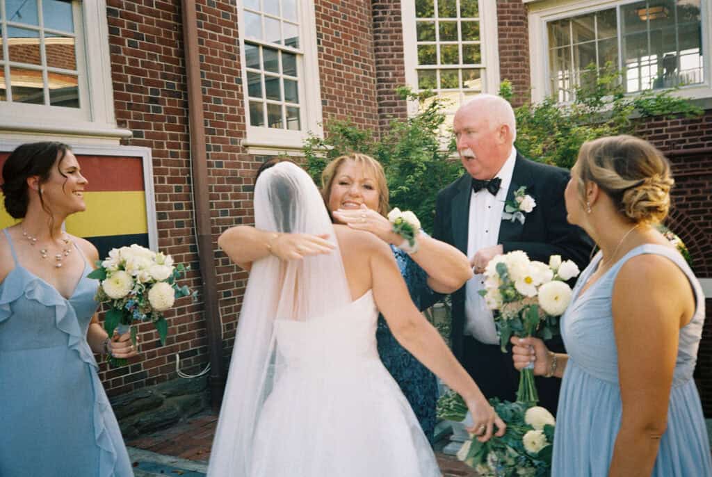 Bride embracing her mother in law while family and bridesmaids in light blue dresses look on outside the brick clubhouse, on film