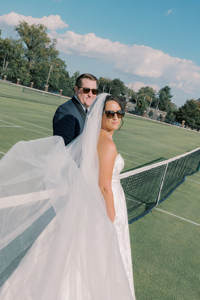 Bride and groom in sunglasses stand back to back by the tennis net, her veil sweeping across the grass court
