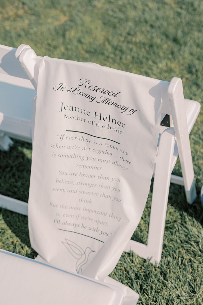 Memorial chair sign reading "In Loving Memory of Jeanne Helner, Mother of the bride" draped over a white ceremony chair on the lawn