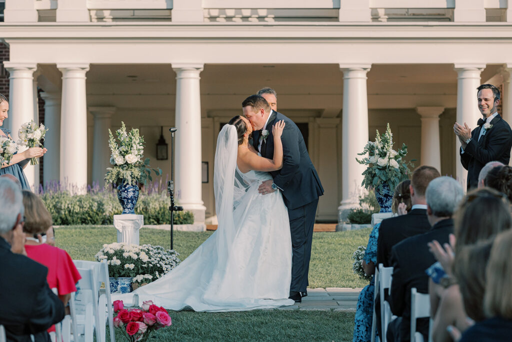 Bride and groom share their first kiss beneath the columned clubhouse portico, framed by blue and white urns of white flowers