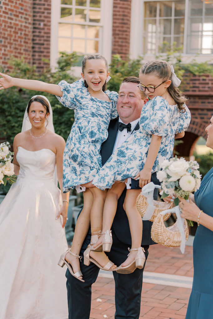 Groom hoists the two flower girls in blue floral dresses on his shoulders as the bride laughs alongside them