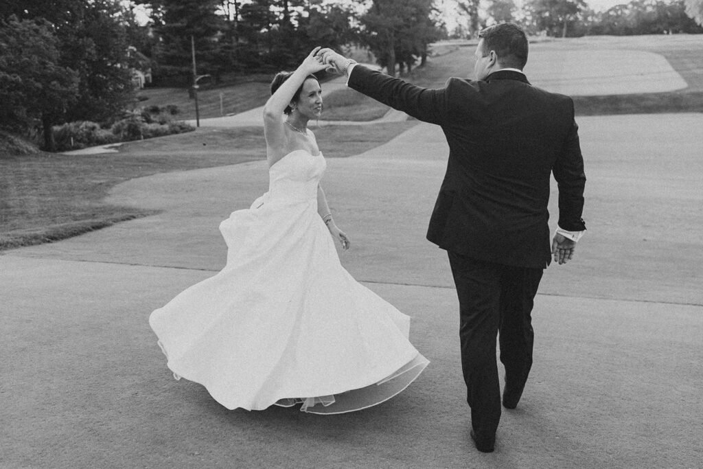 Black-and-white photo of the groom twirling the bride on the golf cart path at sunset