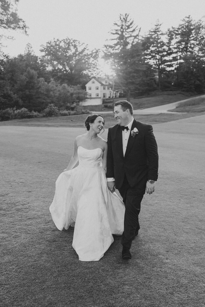 Black-and-white photo of the bride and groom laughing together as they walk the golf course at sunset