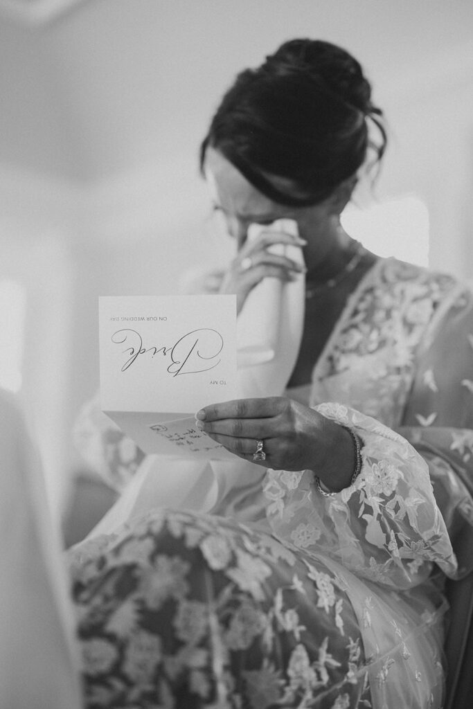 Black-and-white photo of the bride wiping away a tear as she reads a "To My Bride" card in her floral lace robe