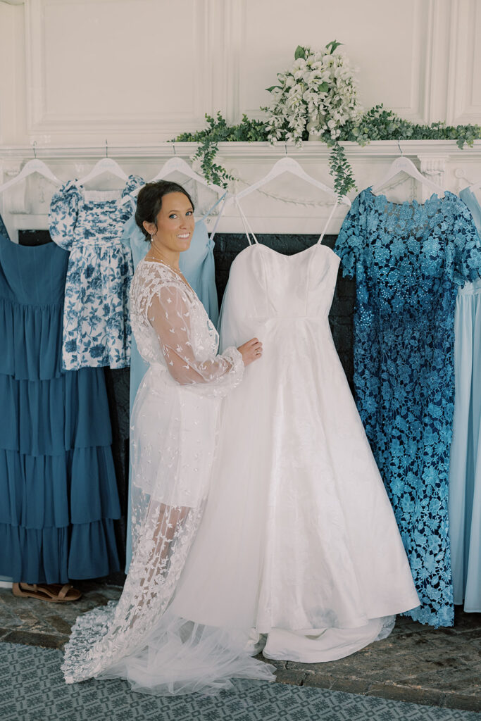 Bride in a lace robe beside her hanging wedding gown, framed by bridesmaid and family dresses in shades of blue