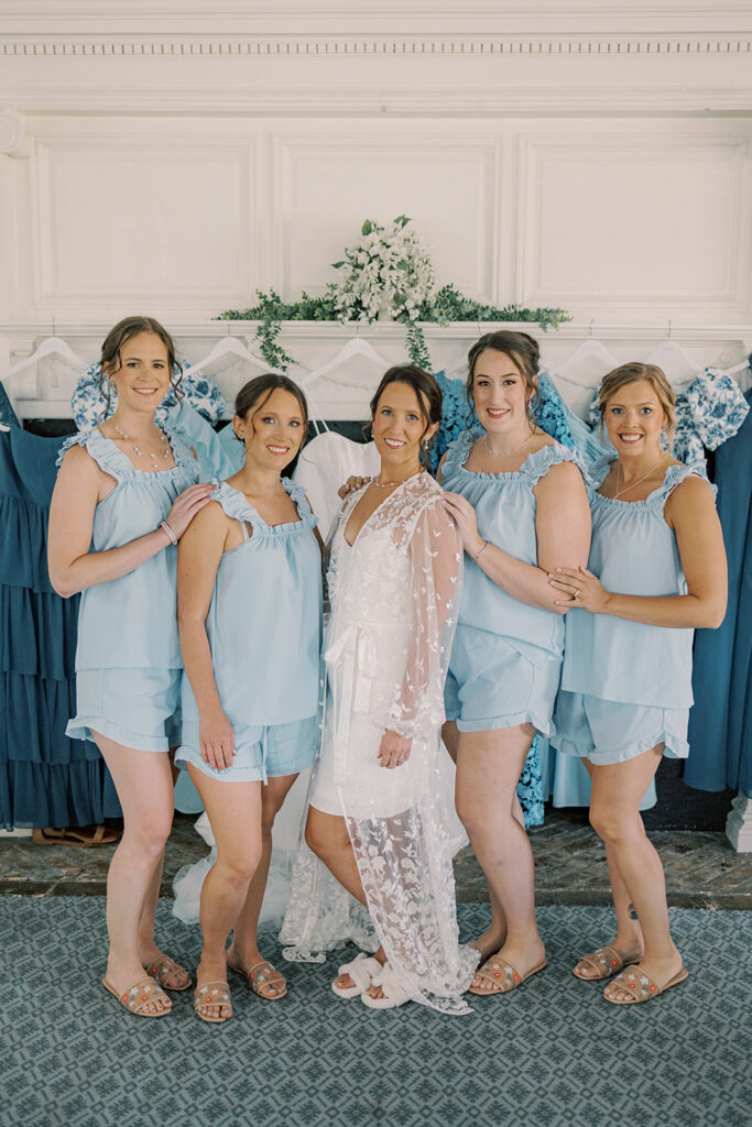 Bride in a lace robe with her four bridesmaids in matching light blue pajamas before a fireplace hung with their blue dresses