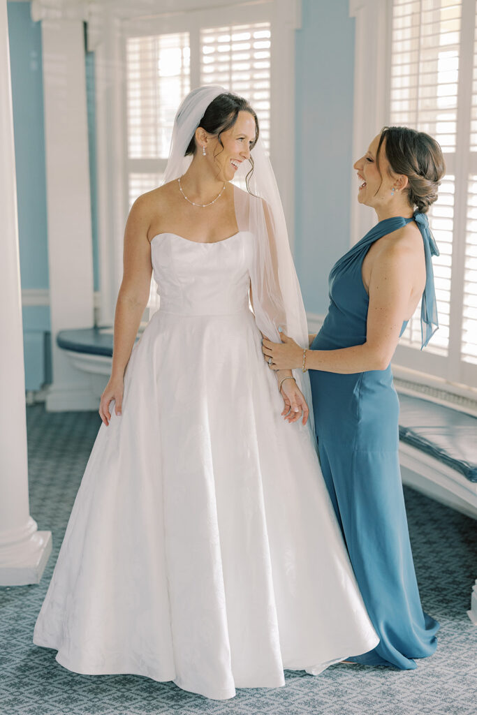 Bride laughing with a bridesmaid in a blue dress as she gets into her gown in the blue suite at the Philadelphia Cricket Club