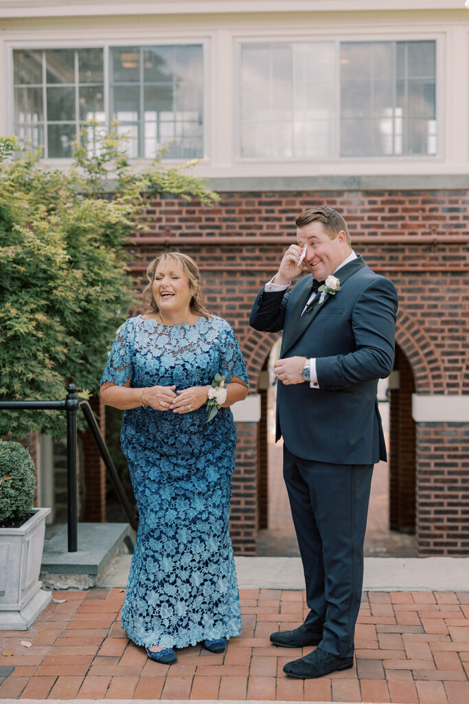The groom wipes away a tear beside his mother in a blue lace gown during a first look on the brick patio at the Philadelphia Cricket Club