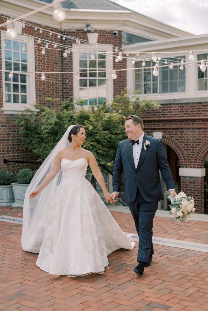Bride and groom hold hands walking beneath café string lights on the brick patio at the Philadelphia Cricket Club
