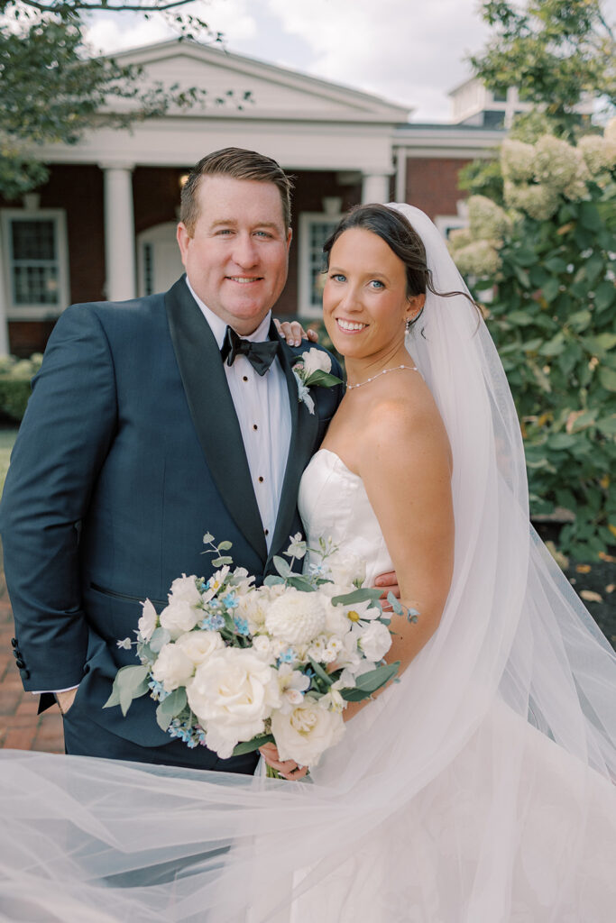 Bride and groom portrait outside the brick clubhouse, the bride holding a white bouquet accented with blue and her veil sweeping around them