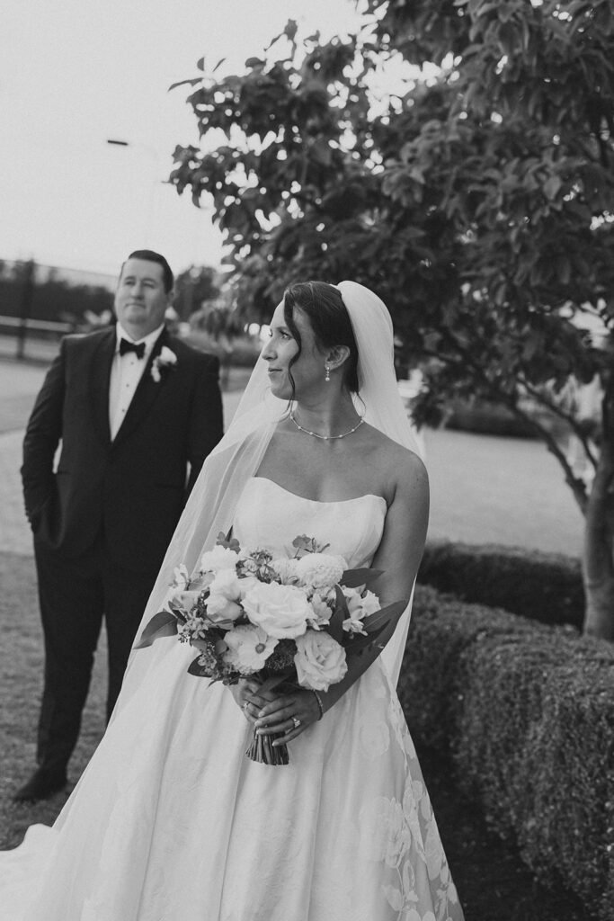 Black-and-white photo of the bride holding her bouquet as the groom stands behind her on the club grounds