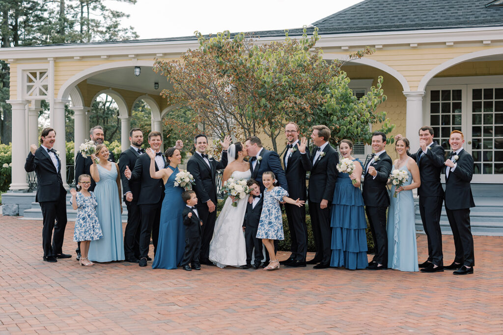 Bride and groom kiss surrounded by their cheering wedding party in shades of blue outside the clubhouse at a blue and white Philadelphia Cricket Club wedding
