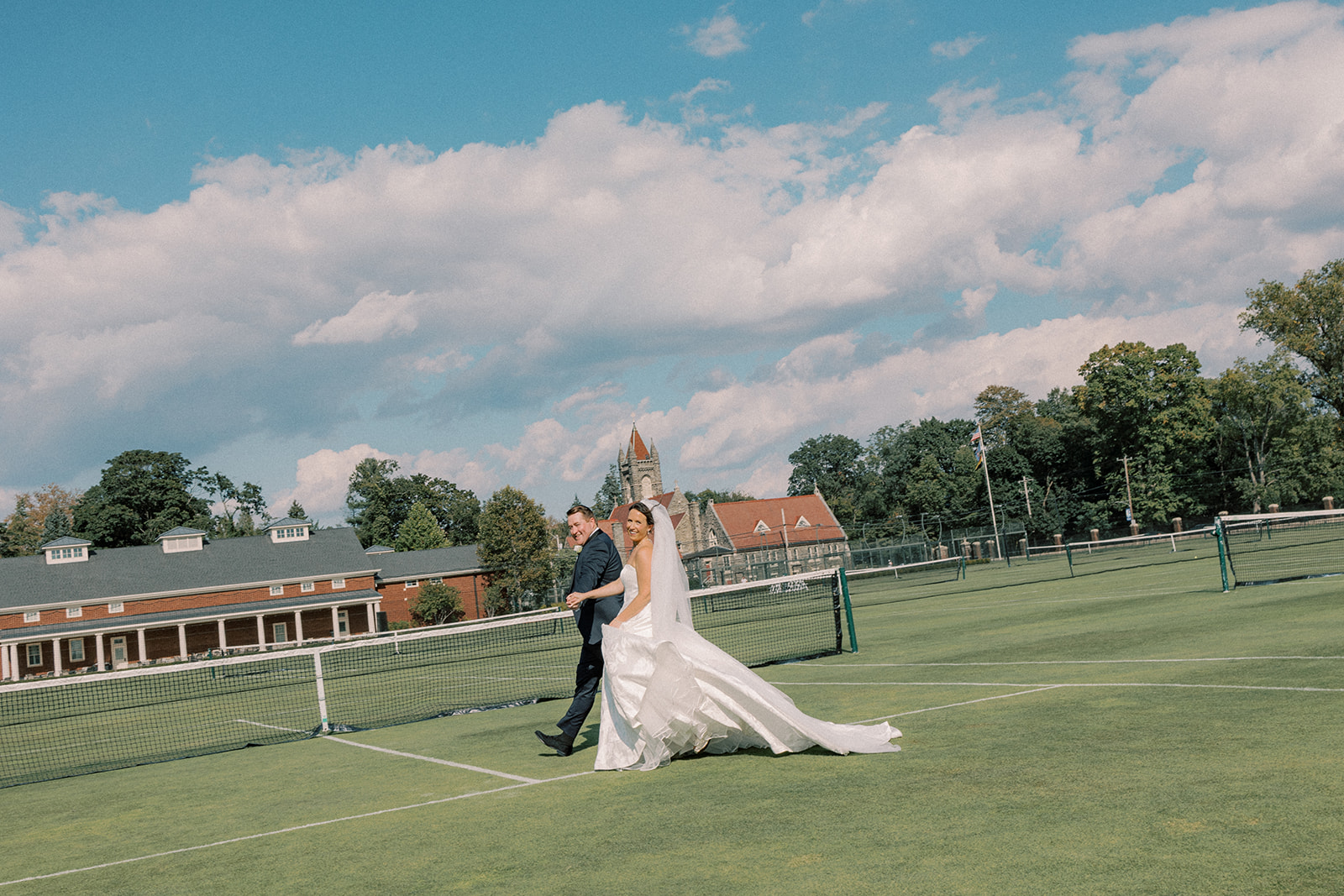 Bride and groom walk across the grass tennis courts at the Philadelphia Cricket Club, the clubhouse spire behind them