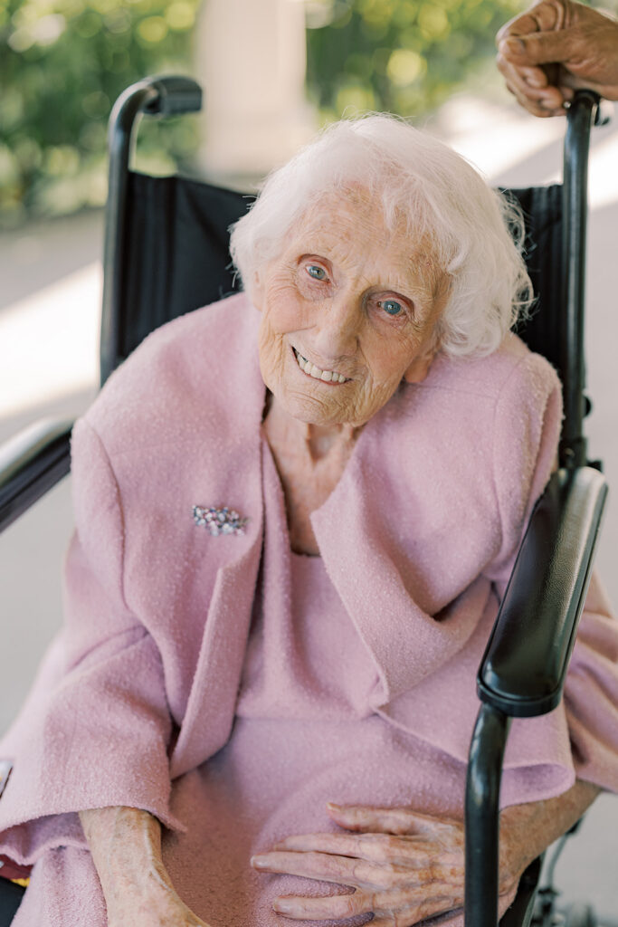 Smiling portrait of the bride's elderly grandmother in a pink suit, seated in her wheelchair on the columned porch