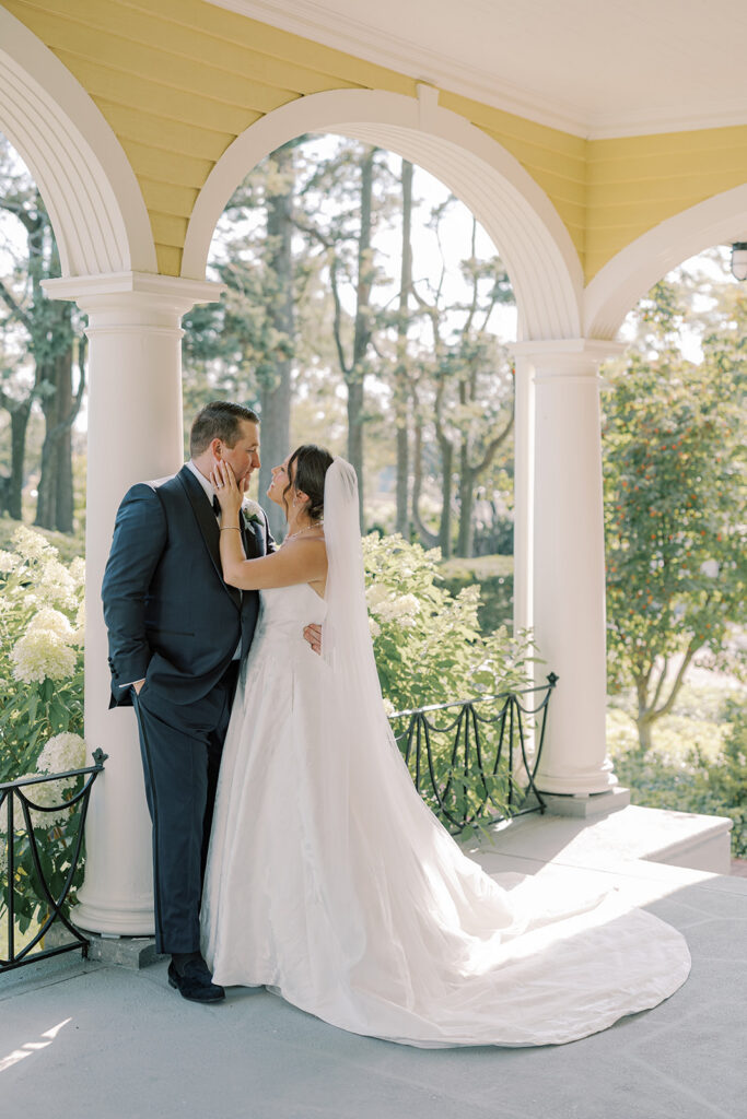 Bride and groom lean in close beneath the columned clubhouse porch, framed by white hydrangeas