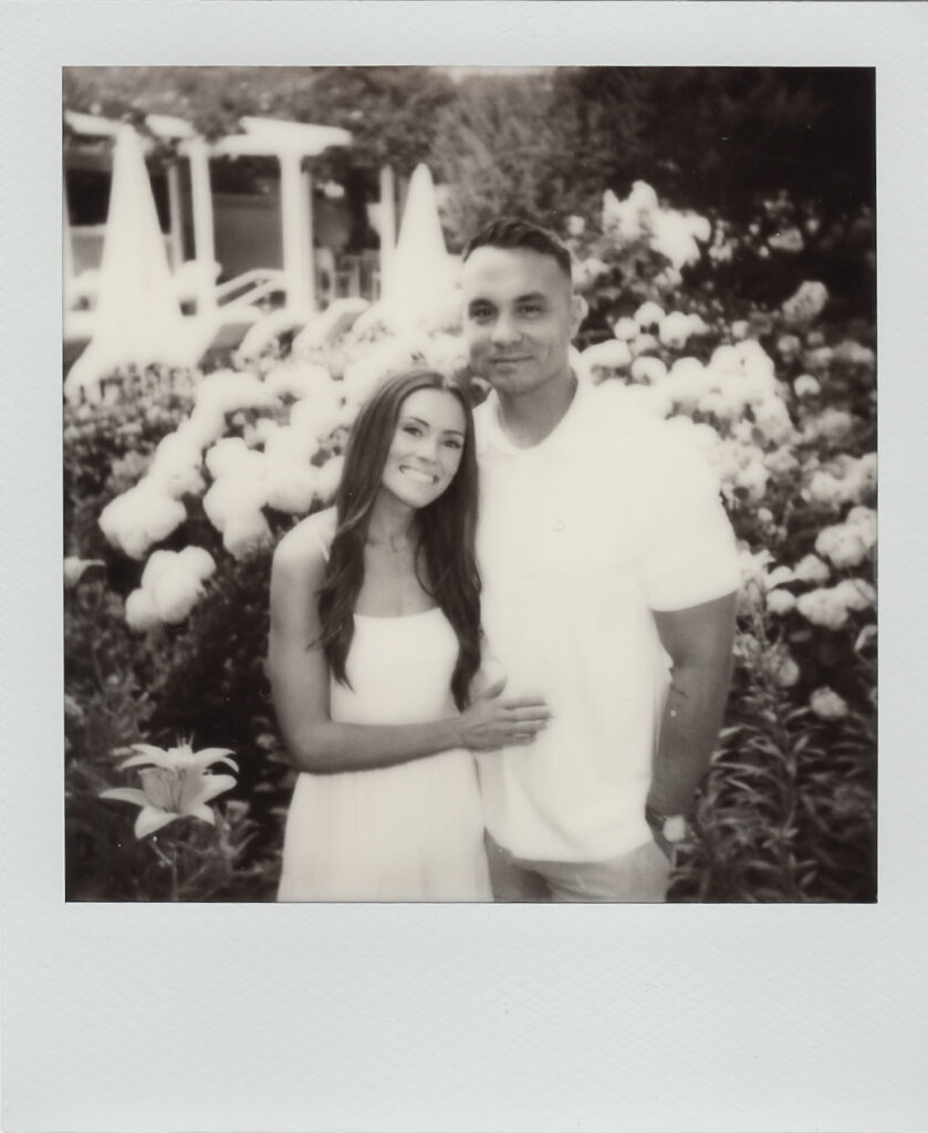 Black-and-white Polaroid of Carolyn and Zach embracing in front of white hydrangeas at Congress Hall during their Cape May engagement session