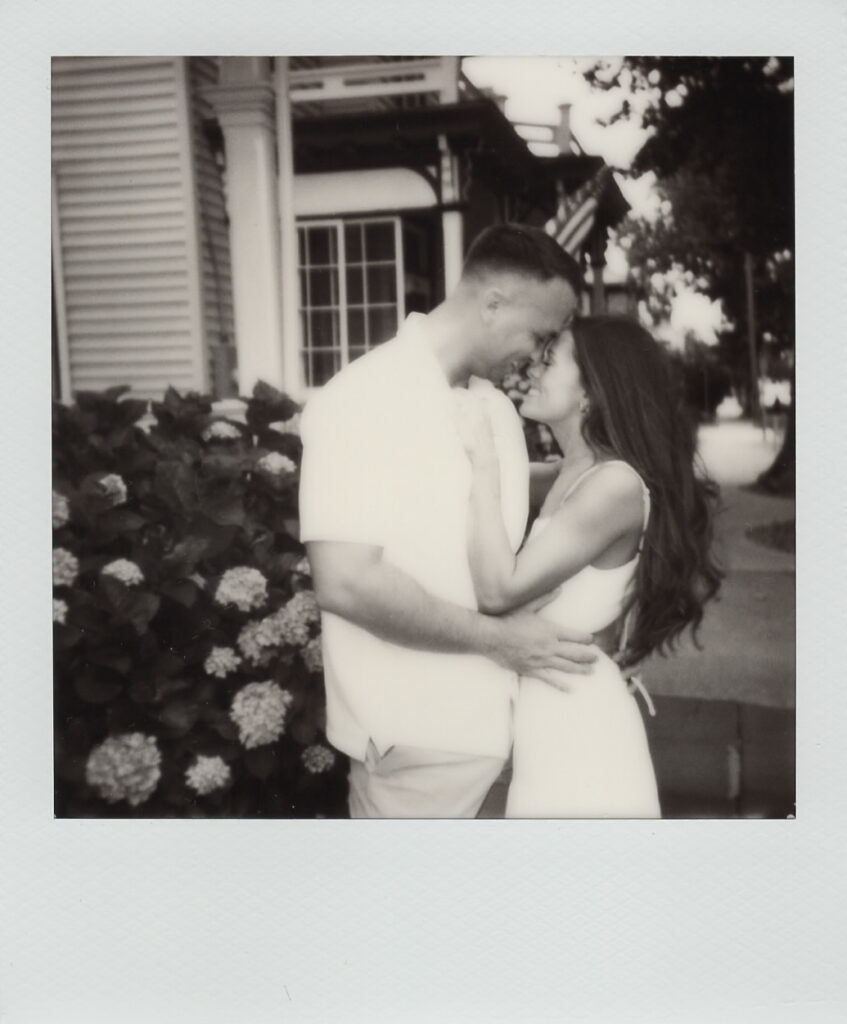 Black-and-white Polaroid of Carolyn and Zach nose to nose beside hydrangea bushes outside a Victorian porch during their Cape May engagement session