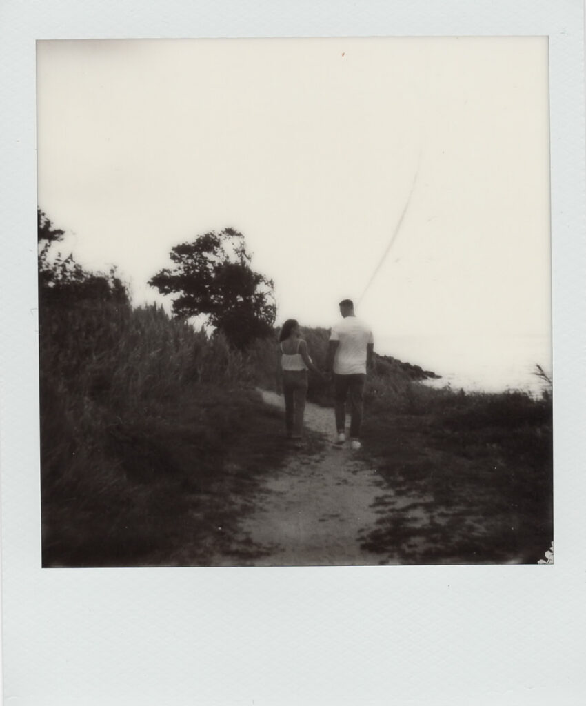 Moody black-and-white Polaroid of Carolyn and Zach walking a coastal path toward the bay at their Cape May engagement session